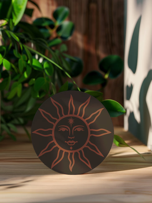 Round ceramic coaster in deep black featuring an intricate burnt-orange sun illustration with a serene, symmetrical face, radiating curved rays, and a delicate star detail on the forehead. The smooth, matte surface enhances the contrast between the dark background and the warm-toned design, creating a celestial, vintage-inspired aesthetic.